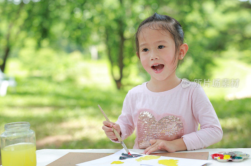 快乐的亚洲儿童女孩坐在一张桌子在一个夏天的花园绘画用画笔。