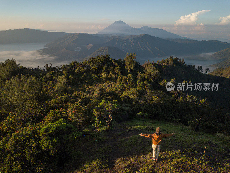 无人机航拍的年轻男子徒步旅行与Bromo火山的观点在印度尼西亚站在山顶手臂张开-人们旅行有趣的冒险概念成功和成就