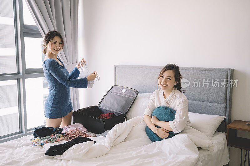 一名亚裔中国女性正在打包行李，而她的姐姐坐在她床上，两人都看着镜头微笑