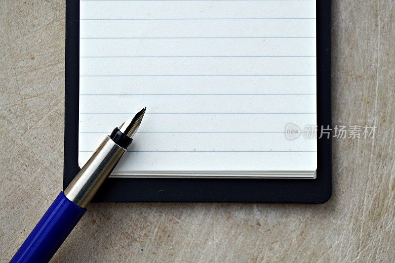 一张水平的照片，一张空白的白色单行页的垂直记事本，黑色封面，上面有一支蓝色和银色的钢笔，背景是木质的米色，陈旧的。