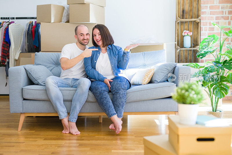 一对年轻的夫妇围坐在沙发上，围坐在搬到新房子的纸箱周围，对着镜头惊讶地微笑着，同时用手和手指示意。