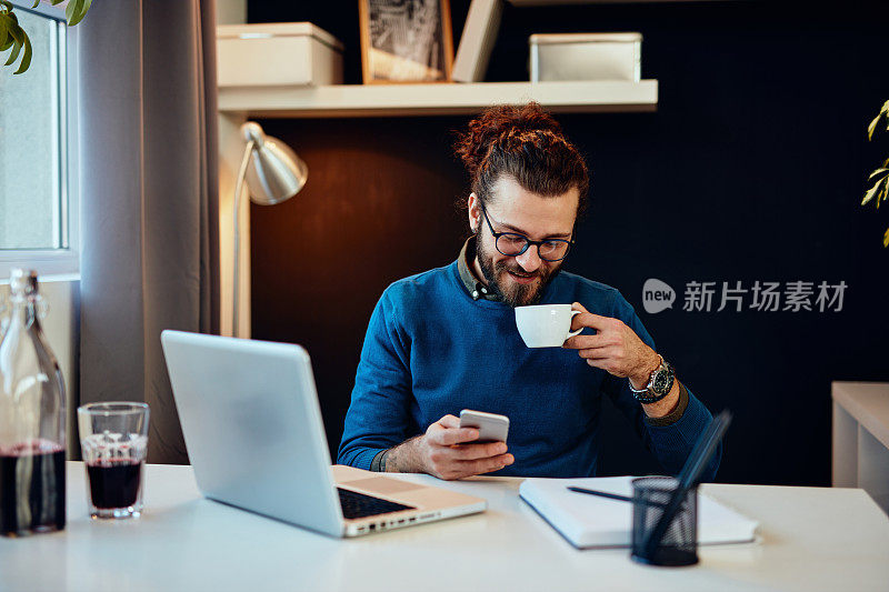 年轻帅气的大胡子嬉皮士坐在他的办公室里，喝着咖啡，用着智能手机。