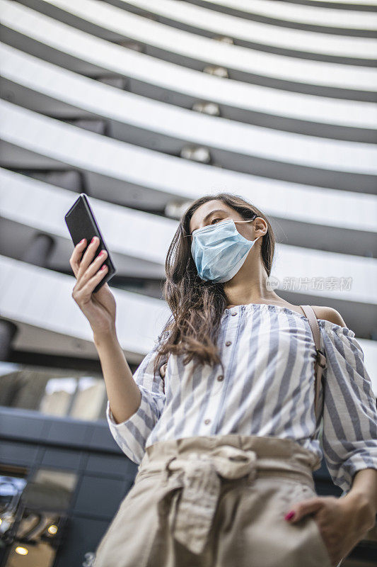 年轻女子站在城市街道上使用智能手机，脸上戴着防护面具。病毒大流行与污染概念。