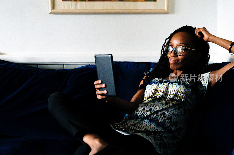 开朗的黑人女性在家里的沙发上用数码平板电脑放松，她平静而满足