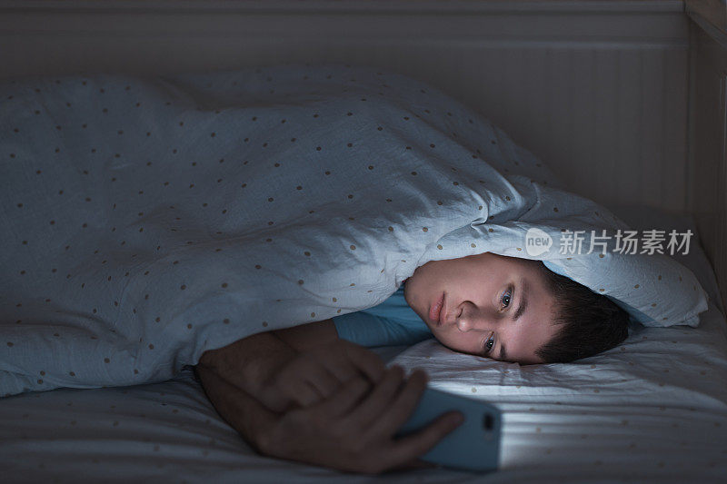 困倦抑郁的少年躺在床上用手机上网。网瘾，智能手机依赖，网络陷阱。