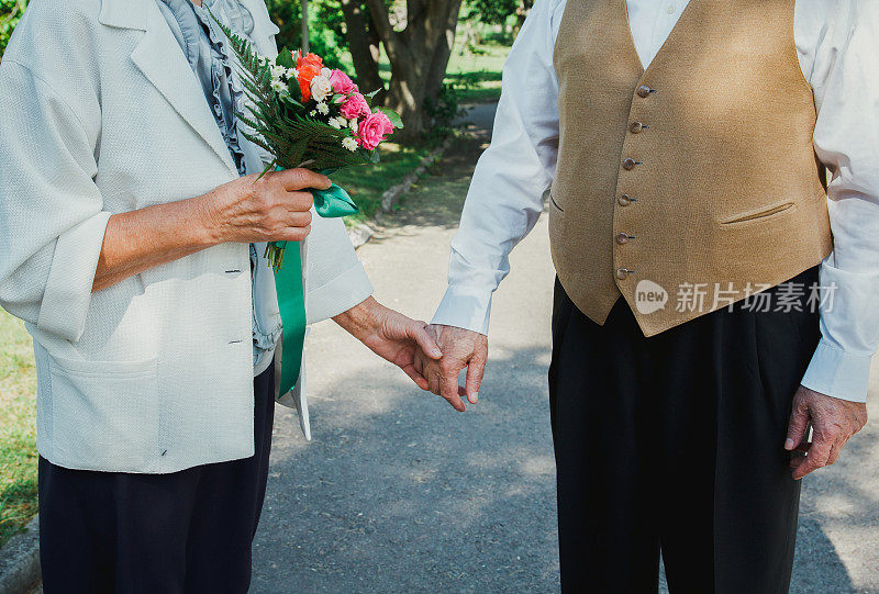 一对老夫妇在绿色的公园里散步。祖母和祖父在他们的金婚纪念日上。50年的爱情故事。奶奶捧着花束，爷爷穿着背心牵着手。