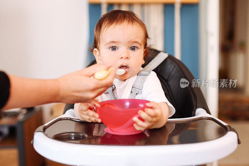 微笑的8个月大男婴坐在家里的高椅子上，妈妈用勺子给他喂固体食物