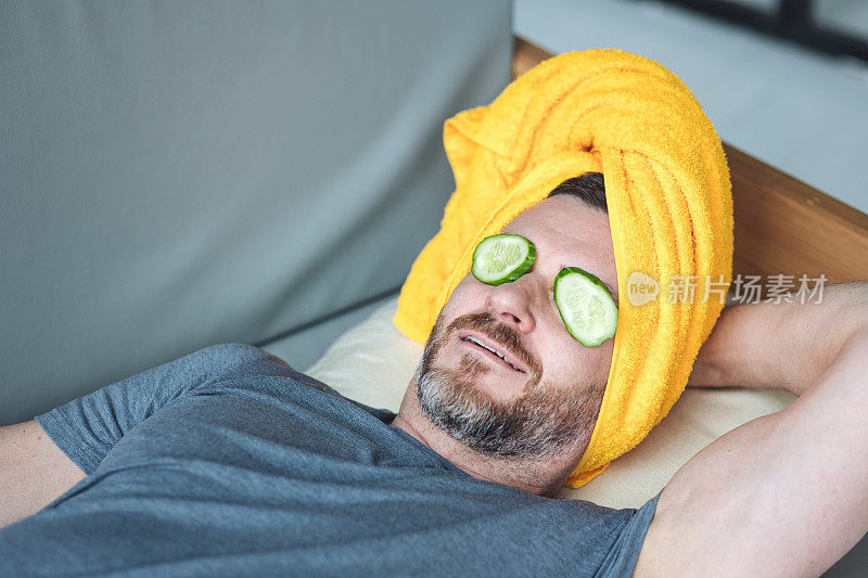 帅哥躺在沙发上，眼睛上放着黄瓜片，头上放着黄色的毛巾。男性自我护理理念，护肤，在家放松。