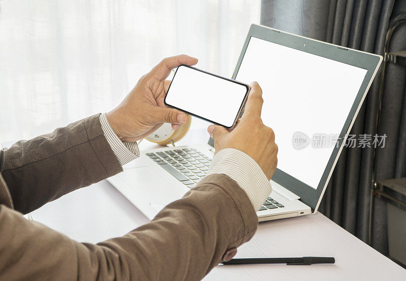 模型形象的商人手拿着智能手机的空白白色屏幕在办公桌和空白白色屏幕笔记本电脑
