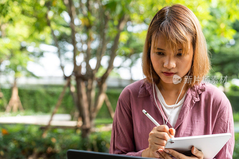 小女孩戴着耳机，用笔记本电脑上网学习，坐在公园里在平板电脑上写字。