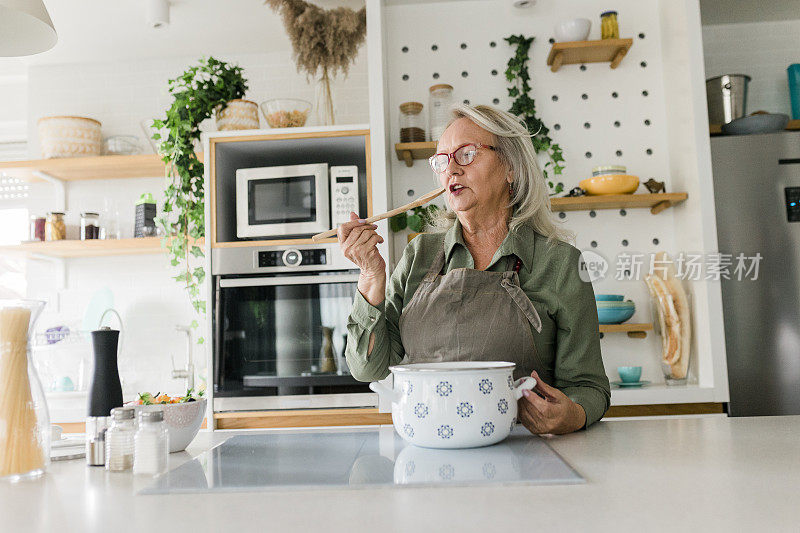 快乐的老年妇女站在厨房柜台前，用勺子搅拌沙拉，同时在炉子上做美味的健康餐。