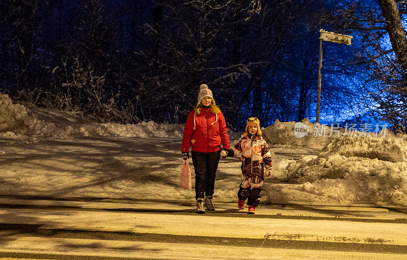 年轻女子，白种人，冬天带着一个七岁的小女孩在街上。这家人晚上在人行横道过马路。