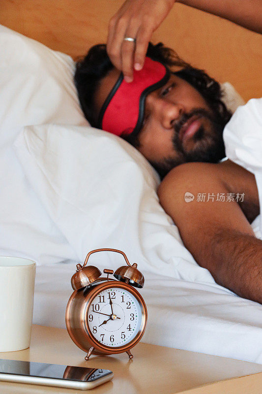 印度男子躺在双人床上，躺在枕头上，从脸上抬起红色睡眠眼罩，看着青铜金属外壳的复古闹钟，双铃，白色表盘的模拟显示时钟坐在床头柜上，白色咖啡杯，智能手机