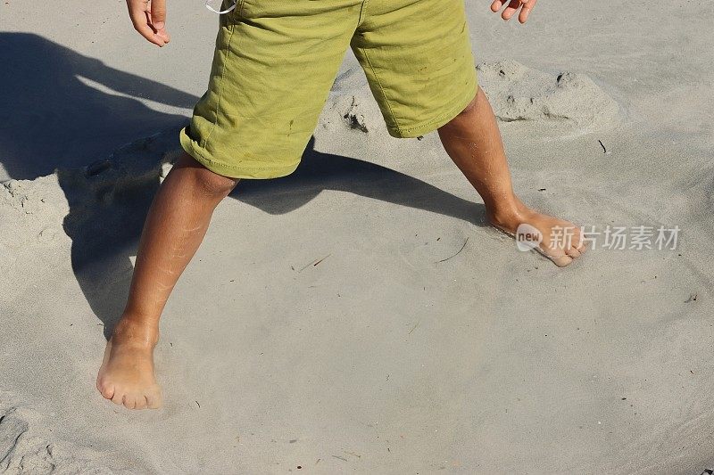 光着脚的孩子的脚和绿色短裤在沙滩上的一滩水里弄湿了。