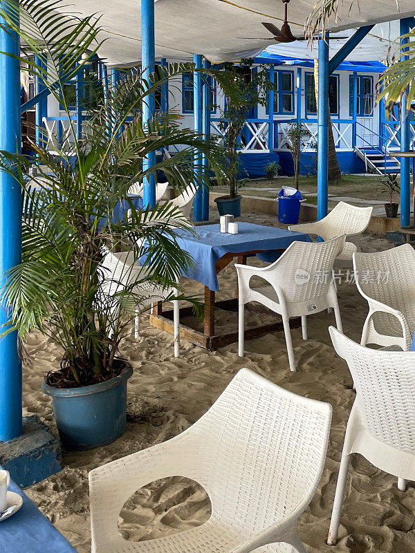 海滩餐厅桌布覆盖的特写图像，金色沙滩上的木制餐桌，遮阳伞下的一组桌子和柳条扶手椅，在印度果阿的Palolem海滩上的户外用餐区，集中在前景