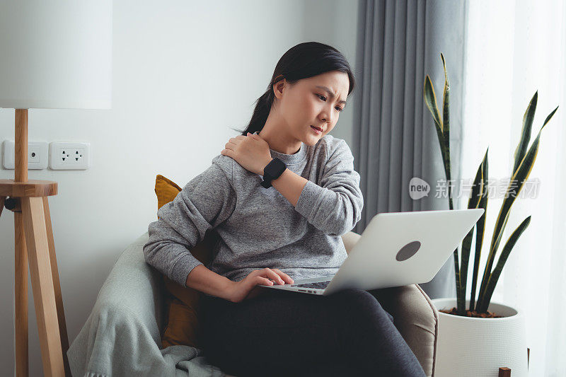 一名亚洲女性坐在家里的客厅里使用笔记本电脑，脖子和肩膀都很痛。