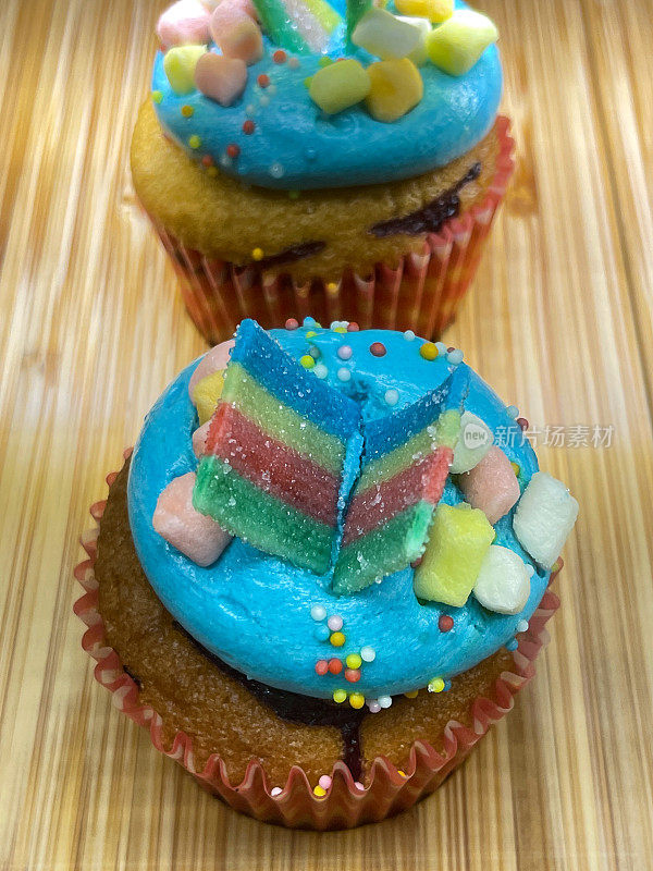特写图片:两个纸杯蛋糕放在蛋糕盒里，装饰着绿松石糖霜和果冻糖果，放在面包店的木架子上，木纹背景，高架视图