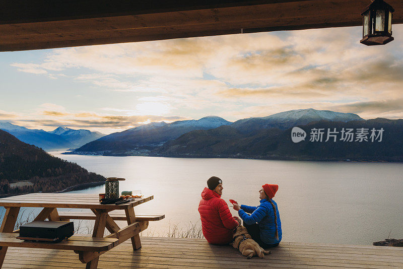 在挪威的日落时分，幸福的夫妇和狗在避难所欣赏冬季峡湾的美景