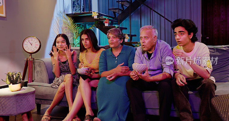 印度幸福延伸的老年家庭坐在沙发上看恐怖惊悚电视，脸上带着恐惧的表情，年长的祖父母，年轻的十几岁的男孩，女孩，拿着碗，一起吃零食爆米花，深夜在家里看电影