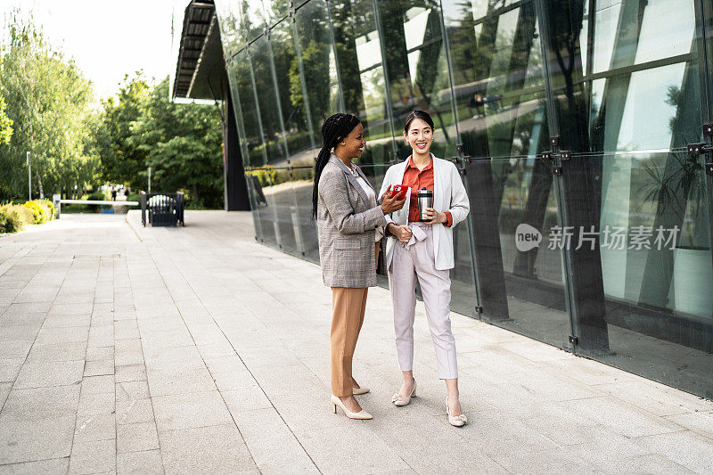 年轻的跨种族商业女同事在办公室外散步和讨论，背景是现代化的建筑