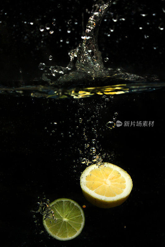 图像切片柠檬和酸橙，柑橘水果落入水中，泡沫和飞溅的水滴在黑色背景下