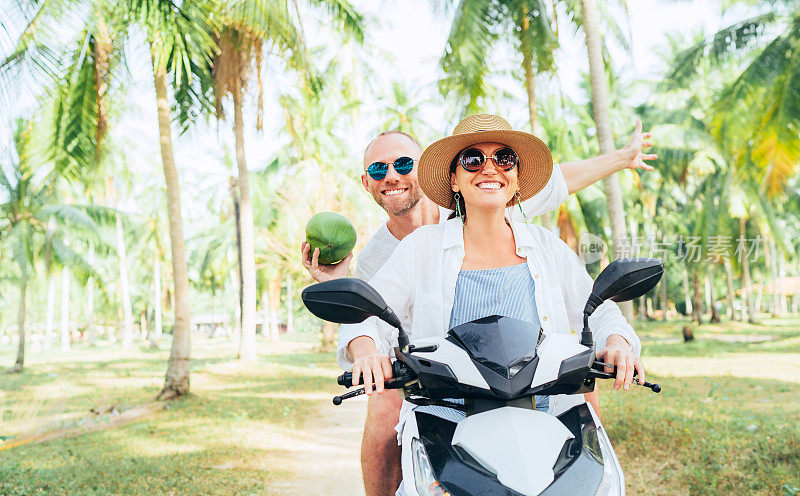 幸福微笑的情侣旅行者骑着摩托车在棕榈树下。手里拿着椰子大笑的男人。热带度假概念形象