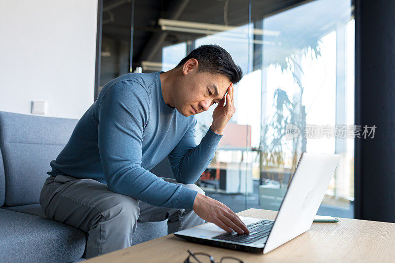 沉思的亚洲人在办公室用笔记本电脑工作，焦虑的商人在现代办公室，沮丧沮丧