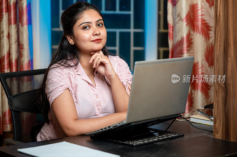 快乐的女人在笔记本电脑上打字，在纸上写字，坐在家里办公室的桌子旁工作。