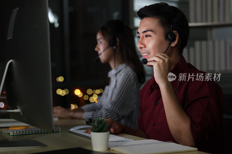 快乐微笑的亚洲人晚上在呼叫中心服务台顾问与队友一起工作，客户服务主管深夜带着麦克风耳机使用电脑支持办公室。