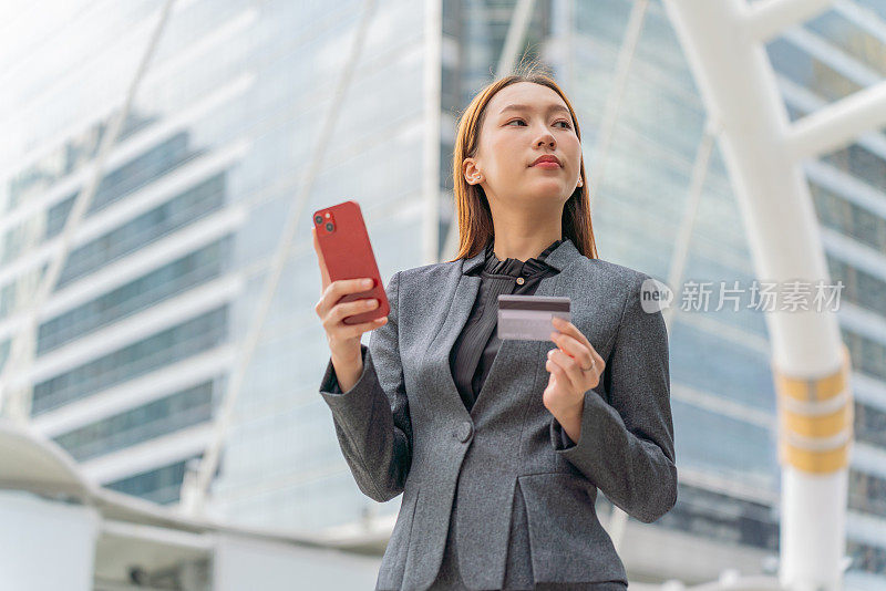 一名年轻的亚洲女商人穿着灰色的办公套装，在商业区用她的智能手机使用信用卡进行在线交易，她微笑着自信