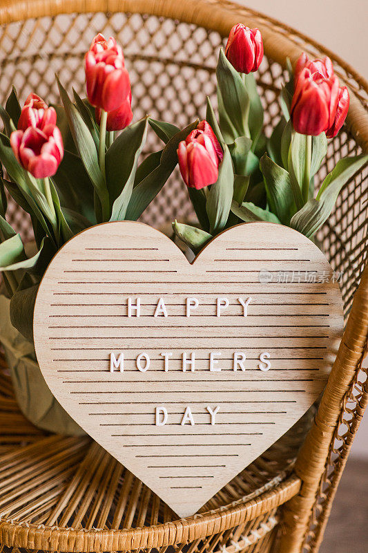 一个心形的字母板，上面写着“母亲节快乐”，明亮的粉红色郁金香盛开，郁郁葱葱的绿色茎和叶子在它后面，在明亮的自然光下，坐在复古的藤椅上
