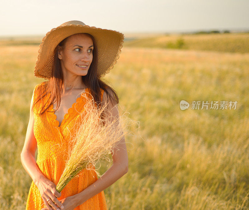 身穿橙色连衣裙，头戴草帽的年轻女子站在夕阳下的田野里