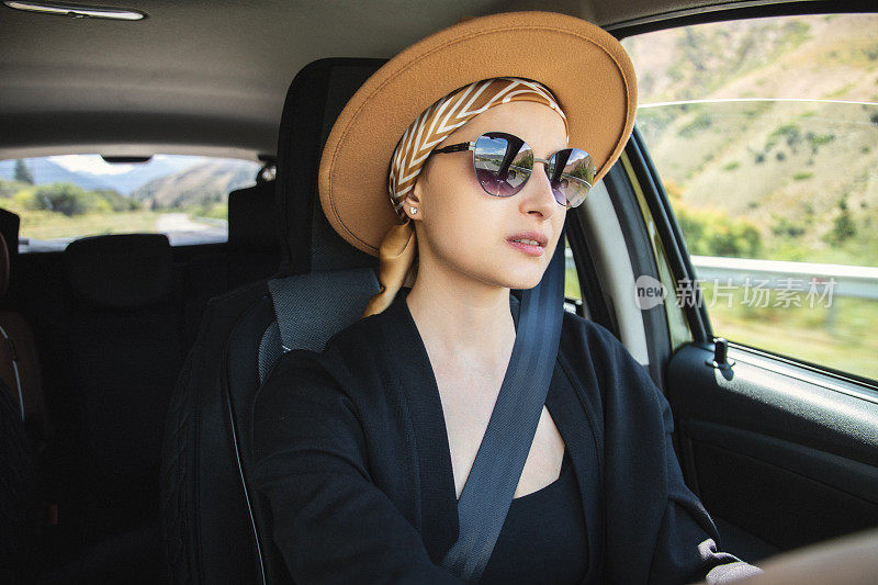 一位年轻的秃顶肿瘤妇女戴着头巾和帽子继续生活，她开着车沿着山路旅行