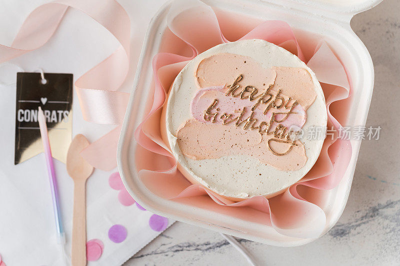 白色礼盒里的小生日蛋糕，还有勺子和蜡烛。时髦的韩式便当蛋糕，上面有生日快乐的题词和粉红色的奶油涂抹。平躺