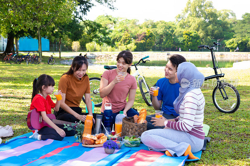 亚洲家庭和朋友一起坐在毯子上，吃着水果和饮料，在公园里享受野餐