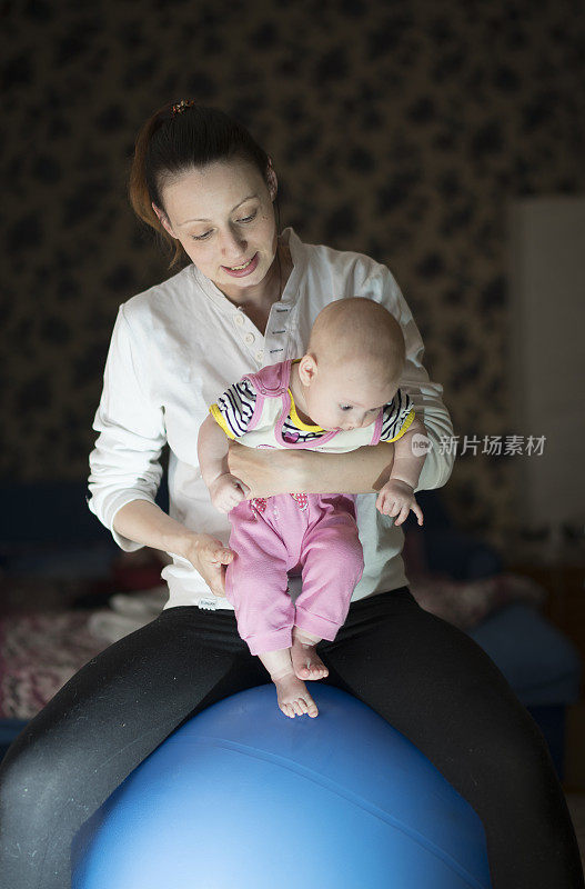 年轻的女人，母亲，坐在健身球上手上抱着6个月大的女儿。