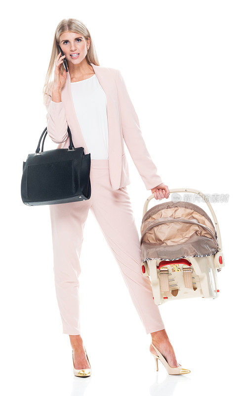 白人婴儿和人类年龄的女商人站在白色背景前，穿着智能休闲装，拿着汽车安全座椅，使用智能手机