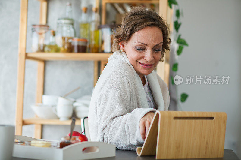 微笑的成熟女性，一边吃早餐，一边坐在厨房柜台前，一边用着平板电脑