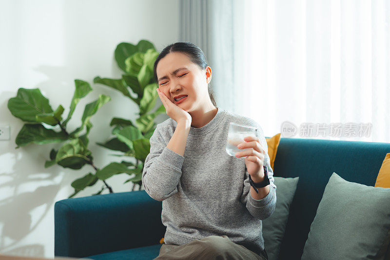 亚洲女性坐在客厅的沙发上，喝了凉水，摸了摸脸颊，就会感到牙齿敏感。