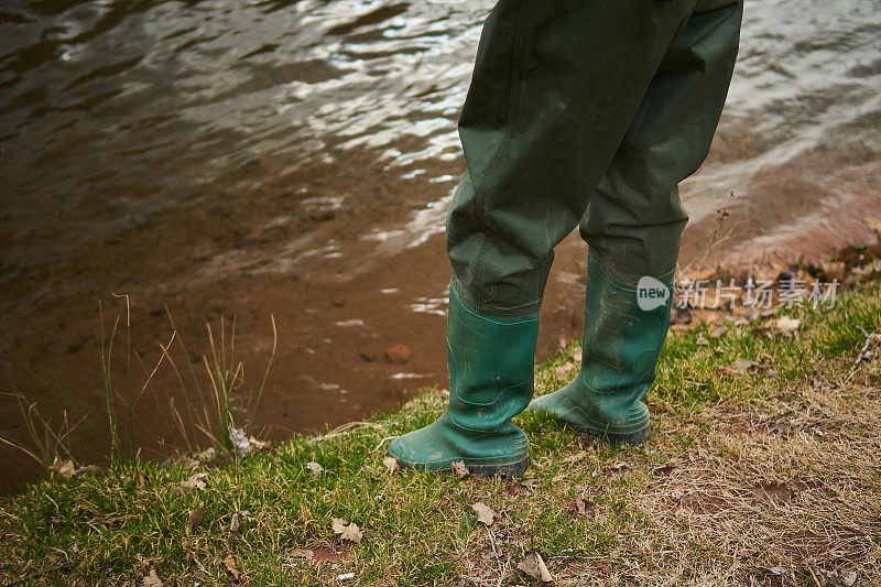 绿色的渔夫靴站在河边泥泞的草地上
