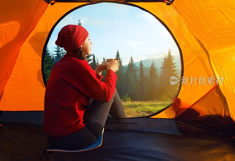 女孩拿着杯子坐在橘黄色的帐篷里看日落