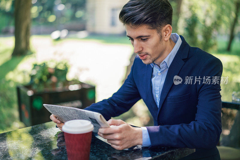 年轻男子穿着西装在外面看报纸喝咖啡