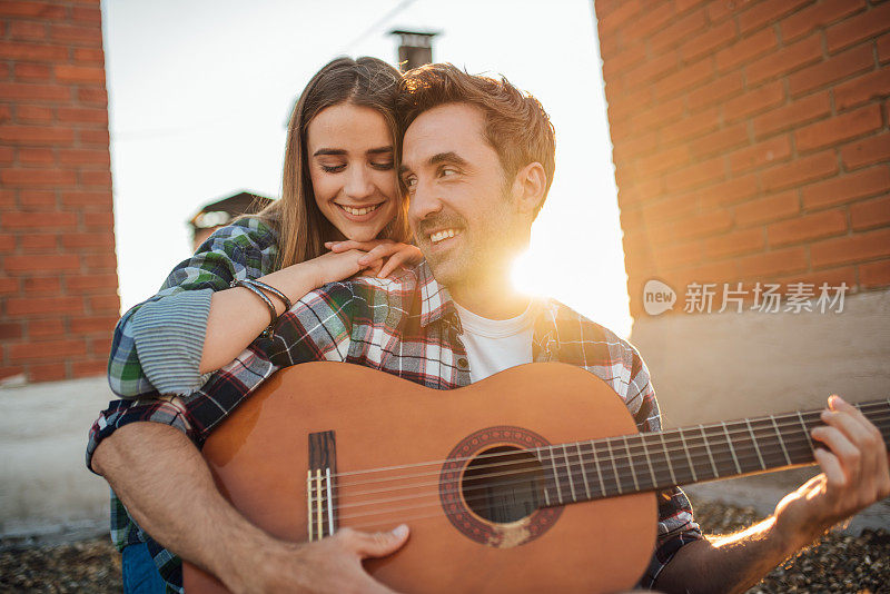 夕阳下，一个年轻人在屋顶上为一个拥抱他的女人弹吉他