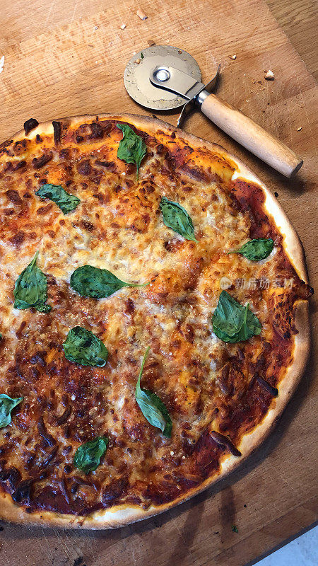 新烤的自制披萨放在木桌上，准备用披萨刀切片。意大利国旗那不勒斯玛格丽塔披萨薄而脆，上面点缀着金色的马苏里拉奶酪，丰富的番茄酱，意大利香草和新鲜的枯萎罗勒叶