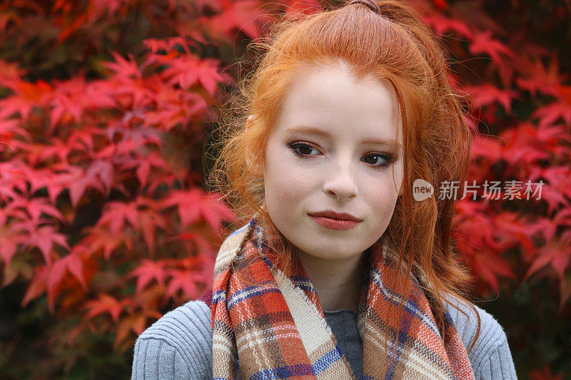美丽的年轻女人的形象少女模特红姜黄色的头发在高马尾和秋天的妆容在花园与红色秋天的颜色，漂亮的青少年看斜面与红色的口红，华丽的自然橙色头发，戴着模糊的叶子围巾