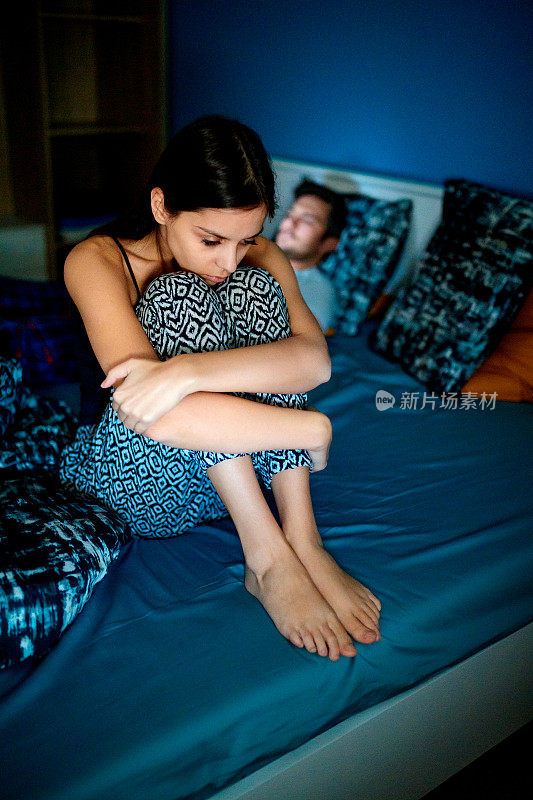 女人坐在床上蜷缩着，丈夫在玩手机