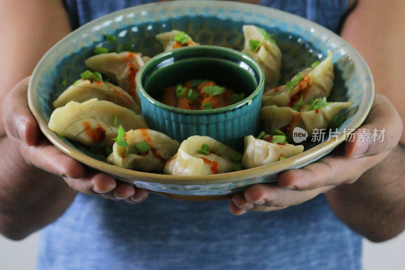 画面中，一名男子端着一盘蒸饺(南亚饺子)，上面是白面粉和盛满鸡肉和杂烩蔬菜的水面团，淋上辣椒油，盘子周围是盛着橘子辣酱的小模子