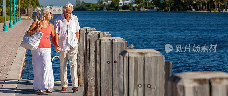 Panorama快乐的老年男女夫妇在热带度假胜地蓝色的海边的码头或木板路上手牵手散步