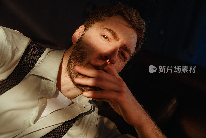 一个留着胡子的男人在黑暗的房间里，直视镜头，抽着烟。