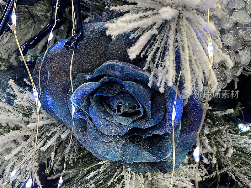 个人人工特写图像，蓝色玫瑰花头装饰品挂在人工圣诞树的树枝上，云杉针上覆盖着人工喷雪，模糊的白色神仙灯背景